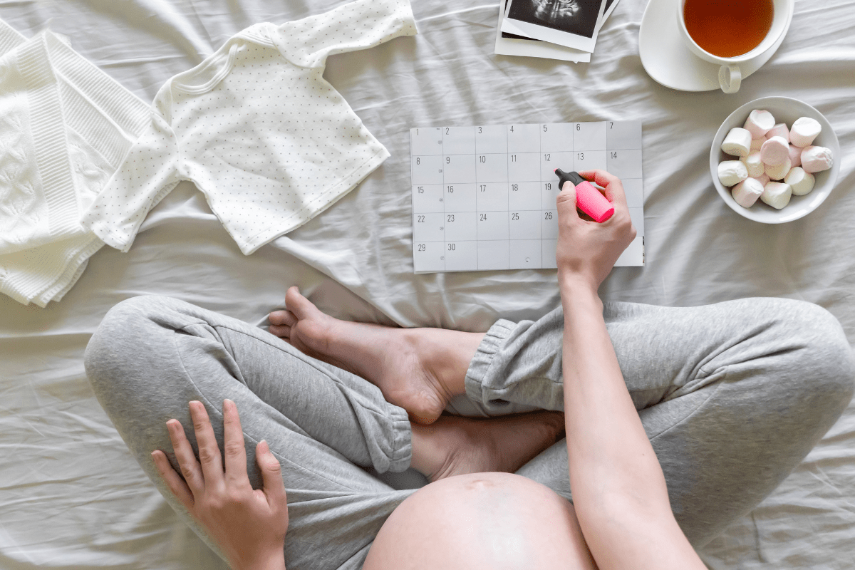 a pregnant woman sitting on a bed with a calendar and a cup of tea