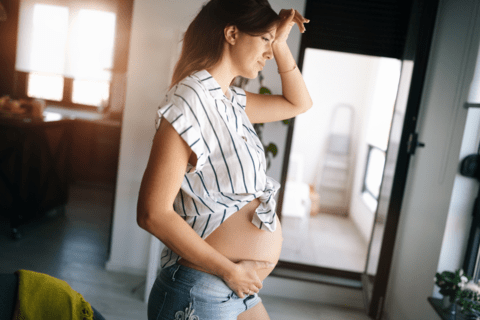 Expecting mother experiencing pregnancy pelvic pain while standing in front of a window