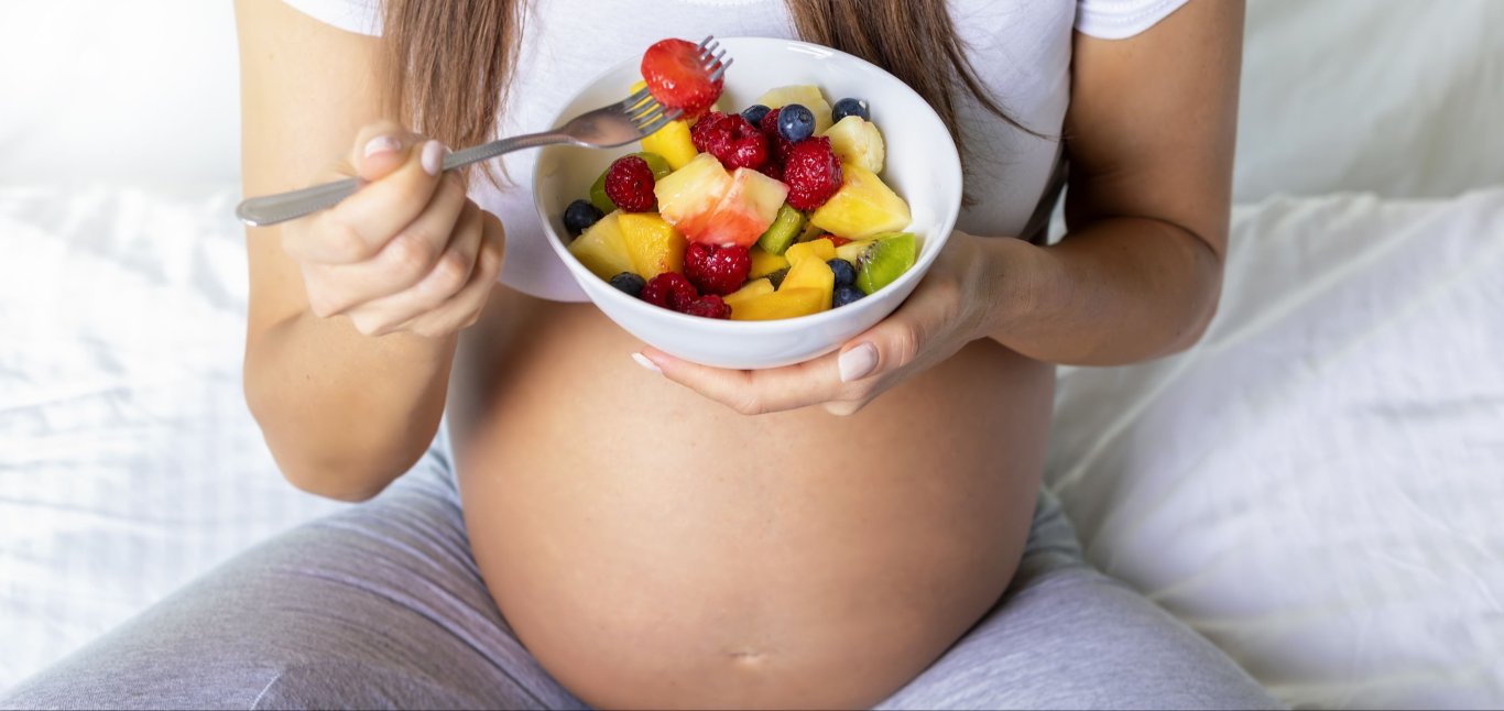a pregnant woman holding a bowl of fruit while sitting in bed