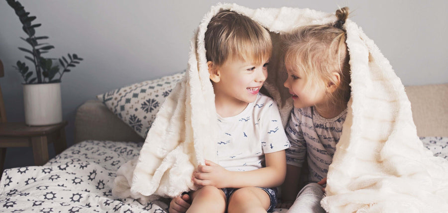 Two smiling children are sitting on a bed, covering themselves with a white blanket