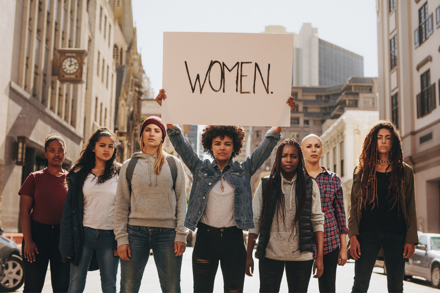 Seven women standing on the street and one of them is holding a sign with “Women.” written on it