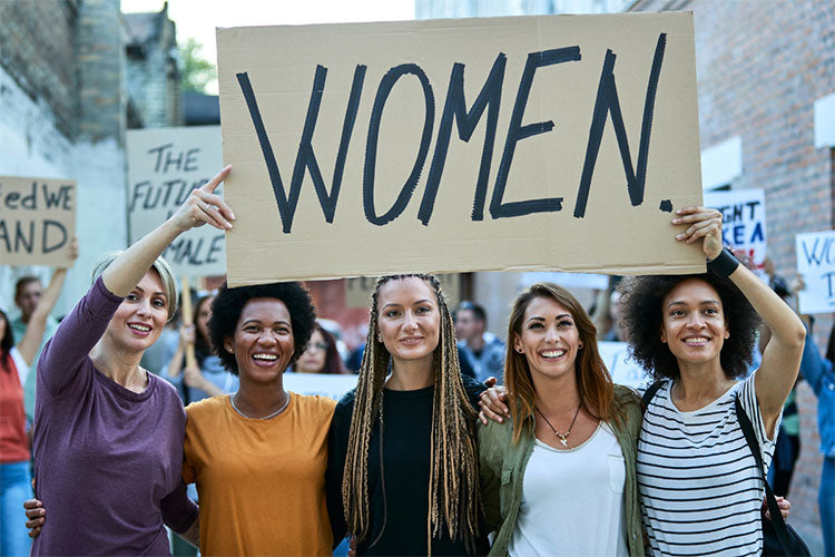 Five smiling women are on the street holding a sign “Women.” with more signs behind them