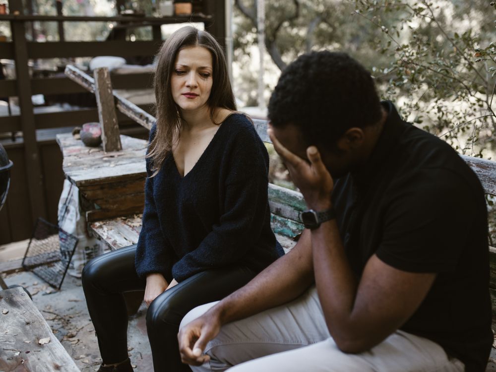 A couple sitting on a bench in a park, with the man appearing sad 