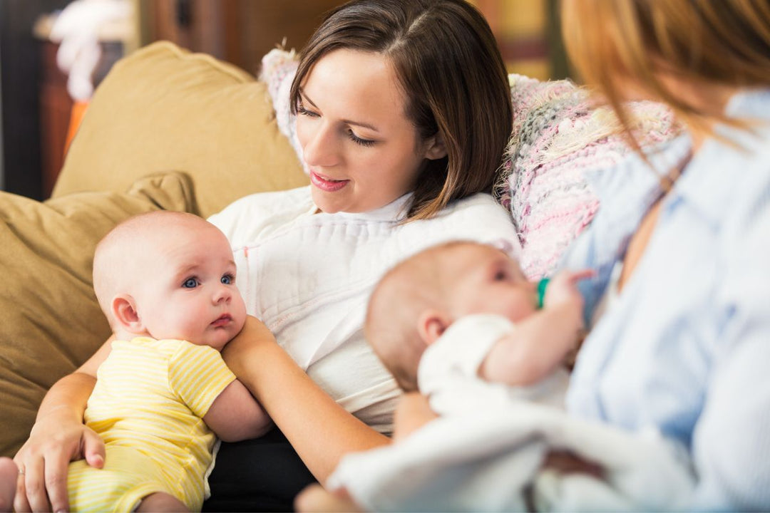Two women sitting on a couch, one breastfeeding her baby while the other sits alongside with her own baby
