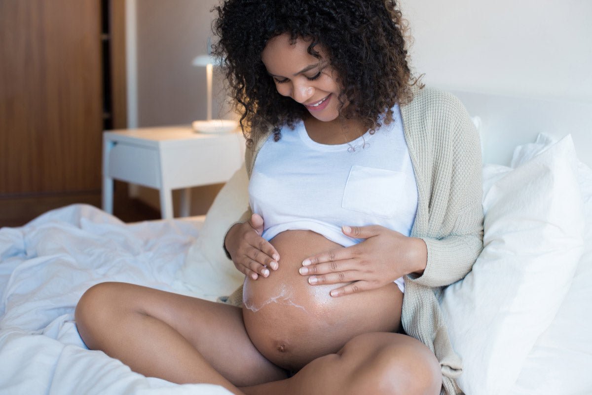 A pregnant woman is sitting on a bed and spreading moisturiser on her stomach