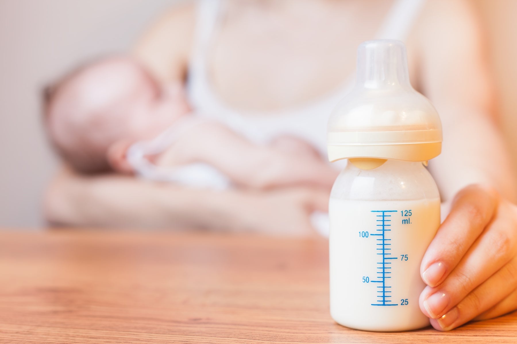  A close-up of a mother’s hand holding a baby bottle filled with formula, while she cradles her baby in the background.