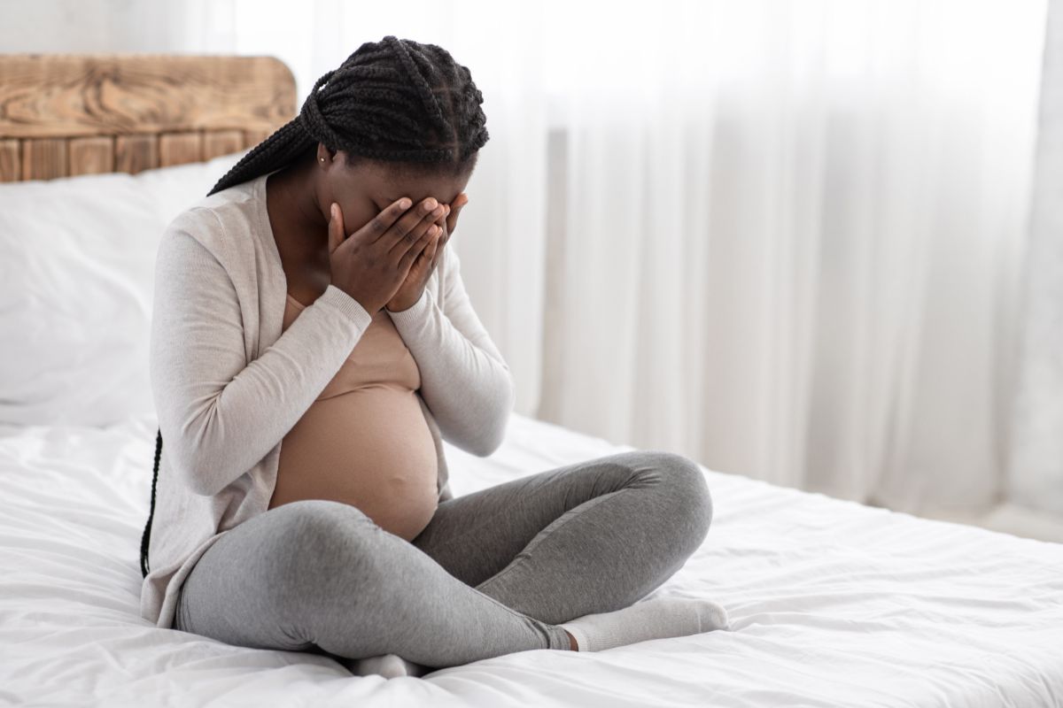 A pregnant woman is seated on a bed and her expression reflects a complex mix of emotions