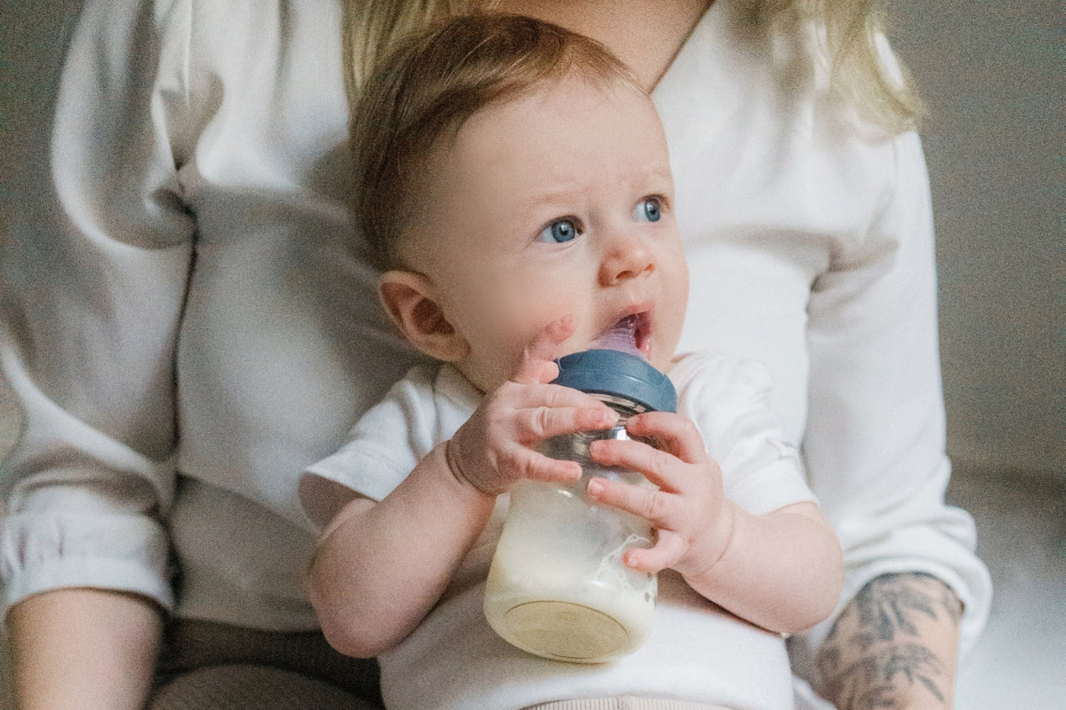 A baby sitting in their mother’s lap, holding a Lola&Lykke NaturalFlow baby bottle, perfect for paced bottle feeding and responsive feeding.