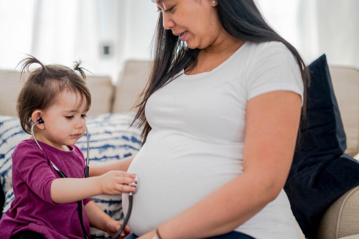 A mum is being pregnant with siblings, while a child is listening to the mum’s stomach with a stethoscope