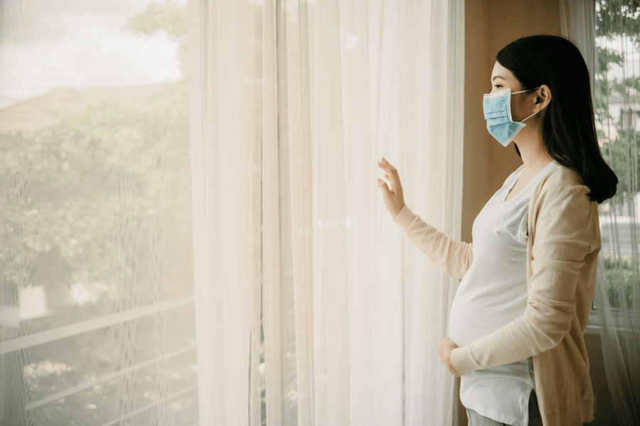 Pregnant woman wearing a face mask and looking outside of a window longingly