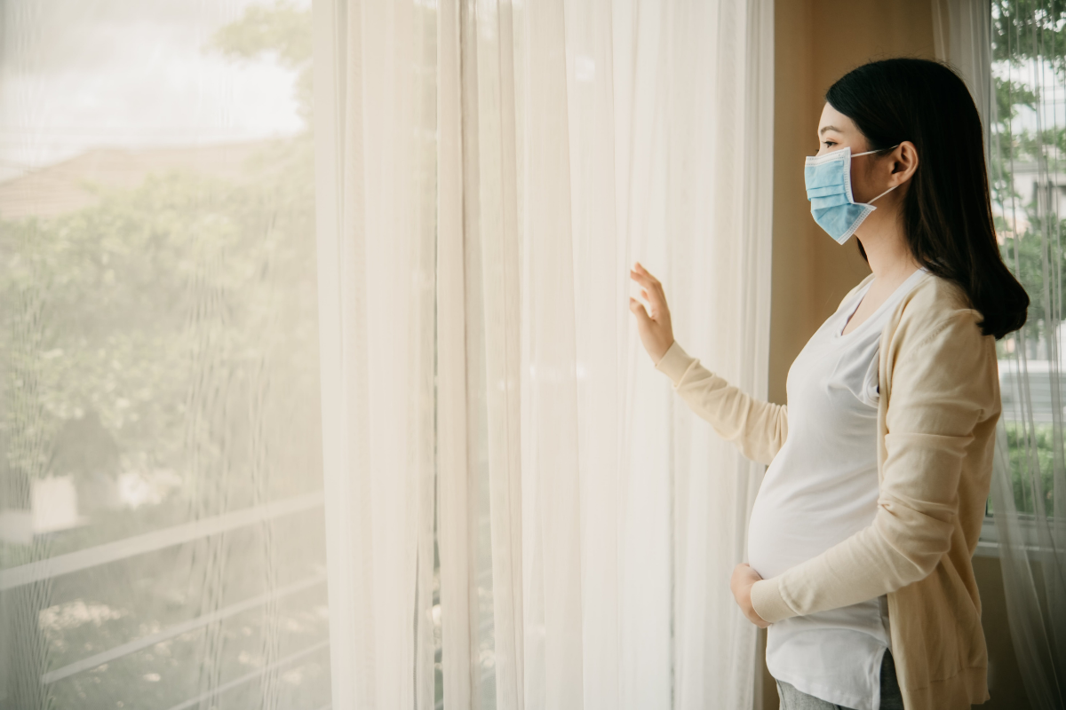 Pregnant woman wearing a face mask and looking outside of a window longingly