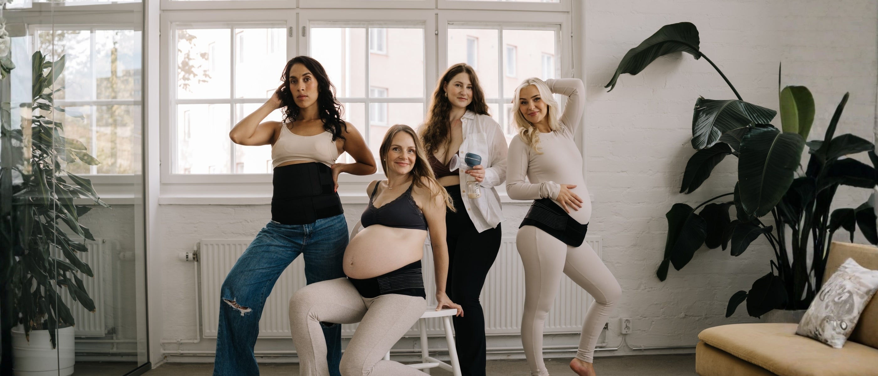 Four women posing together in a bright room with large windows, wearing pregnancy or postpartum bands, with one woman holding a breast pump, showcasing Lola&Lykke maternity and breastfeeding products.