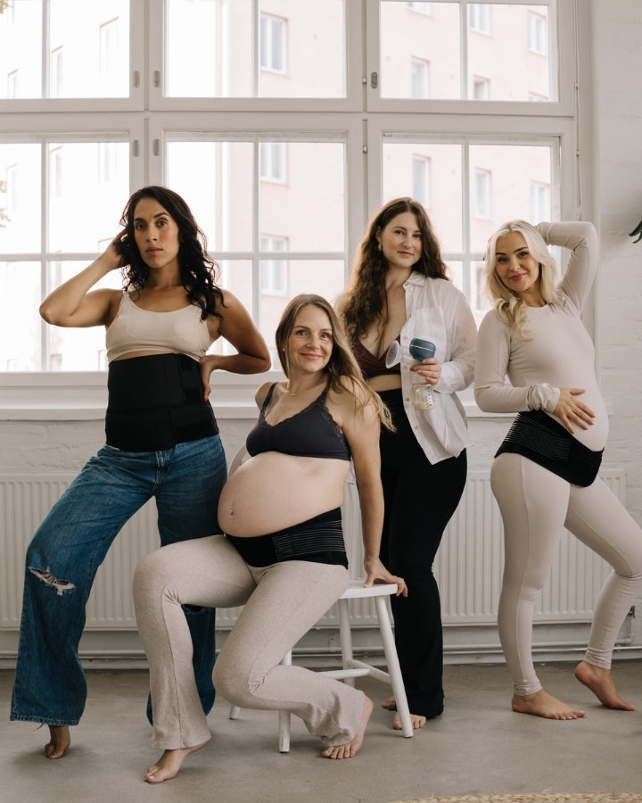 Four women posing together in a bright room with large windows, wearing pregnancy or postpartum bands, with one woman holding a electric breast pump, showcasing Lola&Lykke maternity and breastfeeding products.