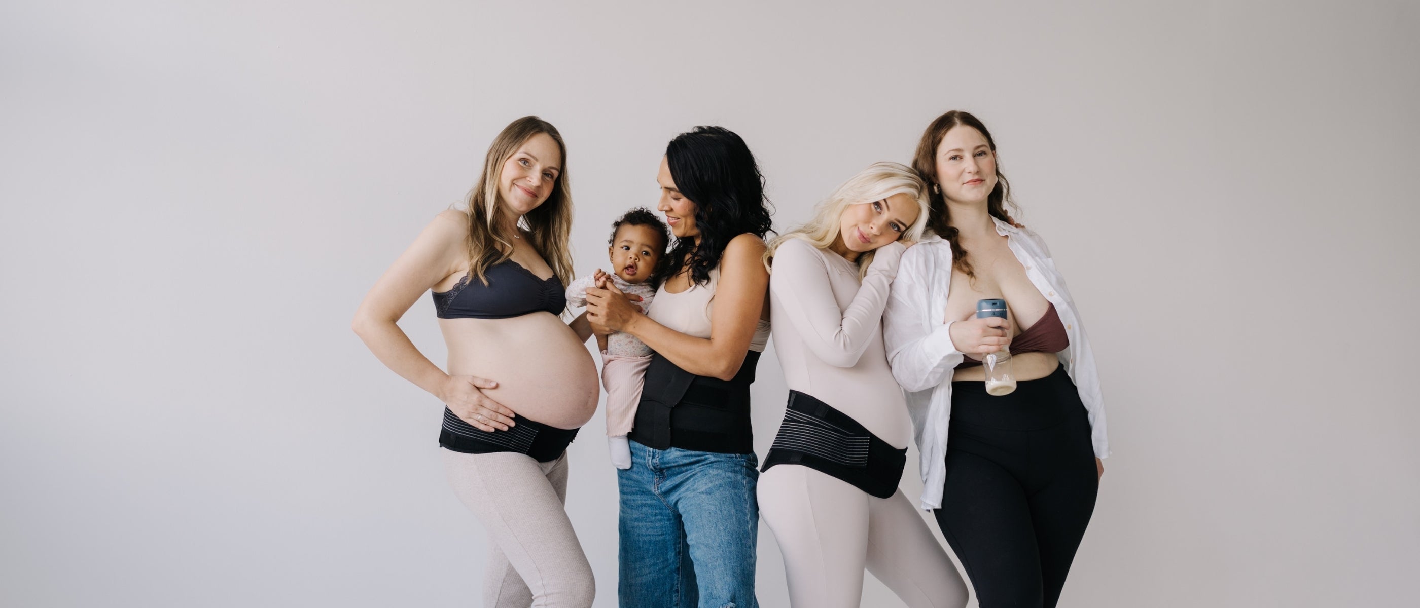 Four women standing together against a white background, one holding her baby, one using a breast pump, all wearing maternity belts, showcasing postpartum and breastfeeding support.