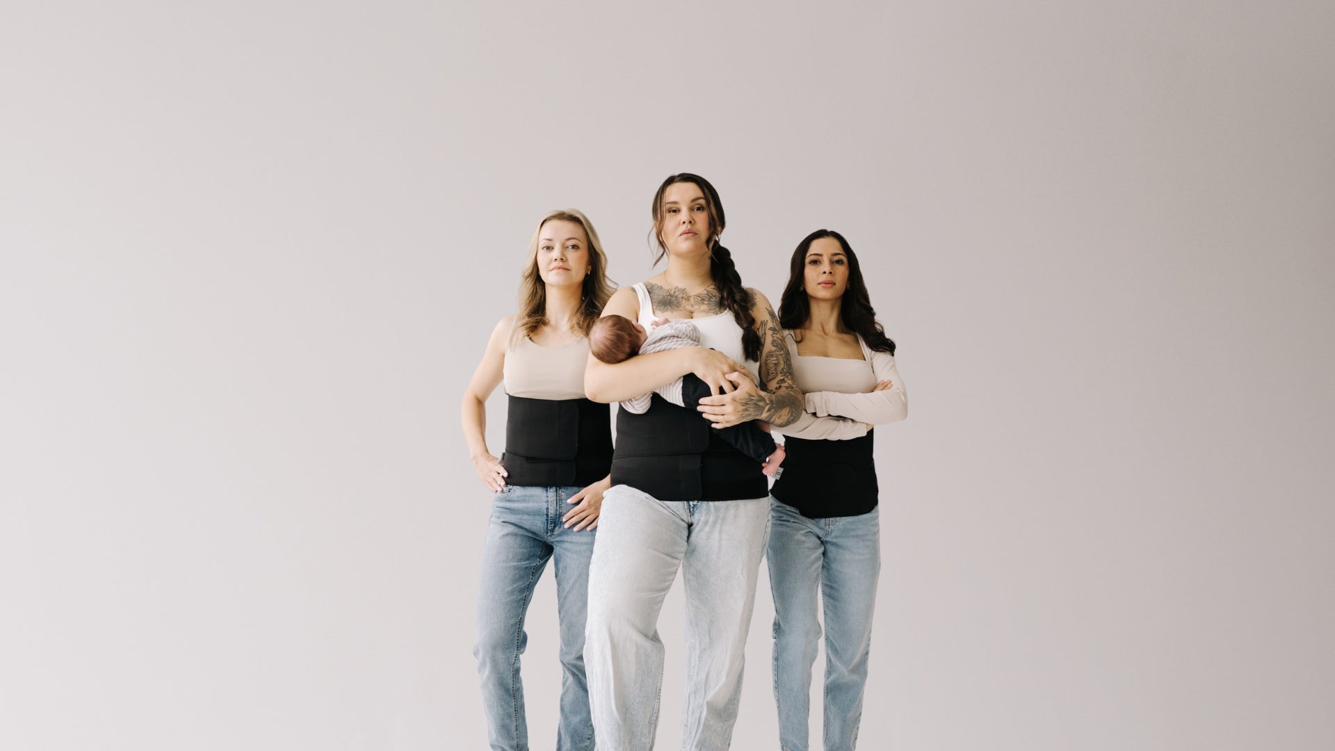 Three women standing side by side against a plain background, all wearing postpartum recovery bands, with one woman holding her baby, illustrating postpartum support.