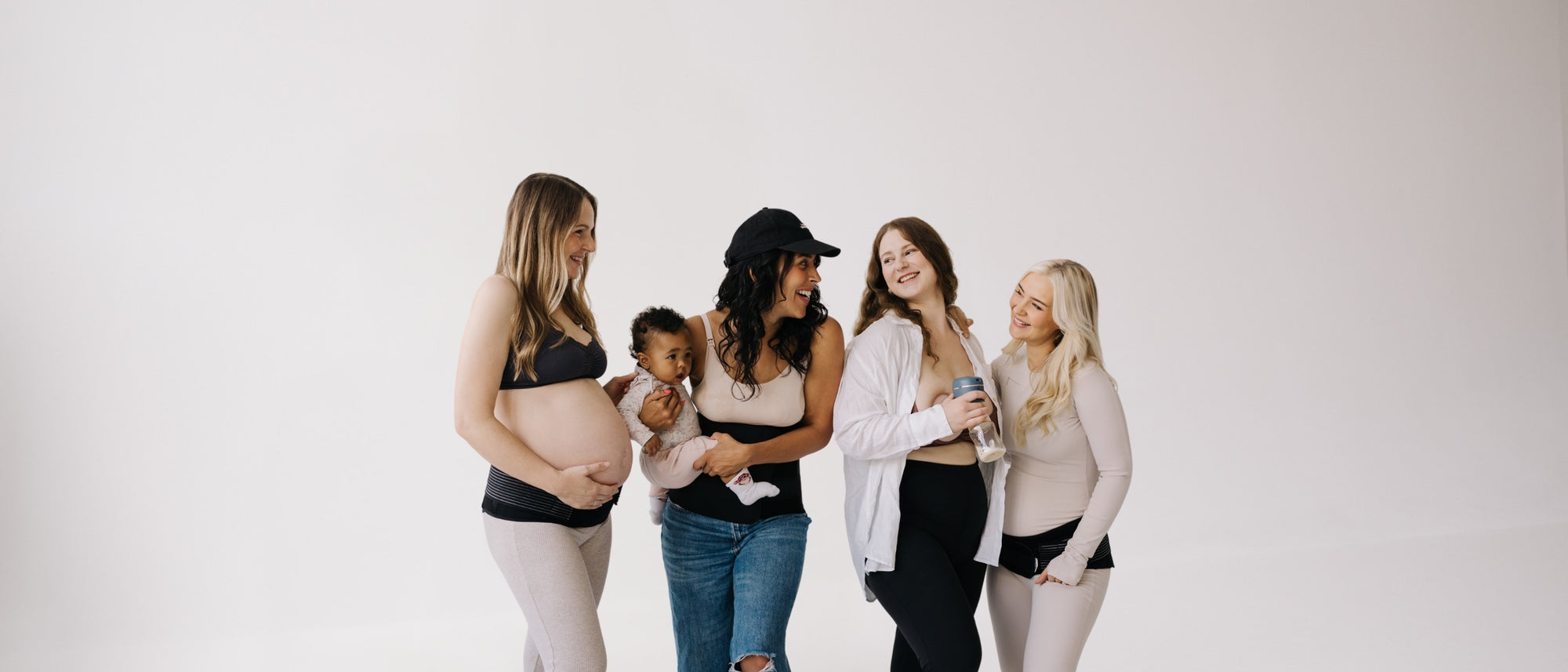 Four women and one baby, including a pregnant woman, standing together against a white background, wearing postpartum or pregnancy bands and holding a electric breast pump, showcasing maternity and breastfeeding products.